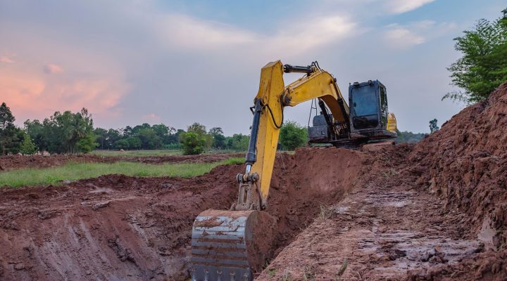 Heavy-Duty Excavator Digging a Trench at Sunset A yellow excavator digging a deep trench in red clay soil against a backdrop of a colorful sunset sky and distant trees.