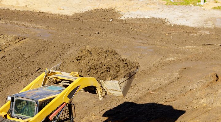 Skid Steer with Grading Bucket for Precision Land Leveling A yellow skid steer loader using a grading bucket to move and level soil on a construction site, casting a sharp shadow in the bright sun.