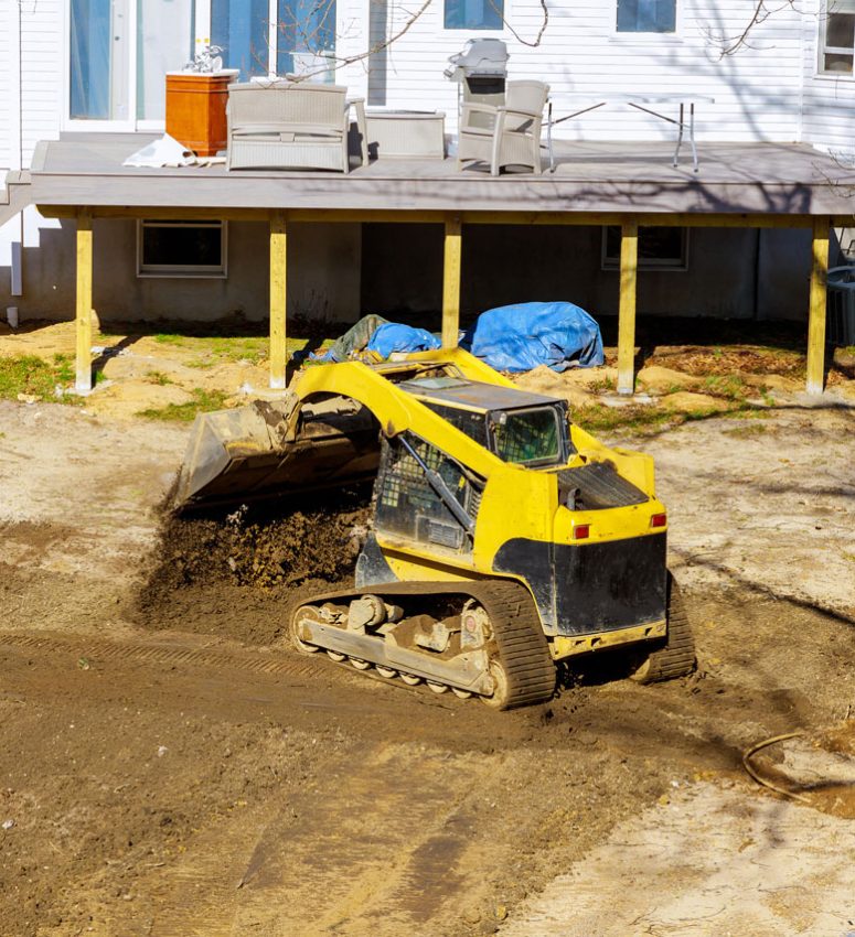 Backyard Grading and Landscaping Project with Skid Steer A yellow compact track loader moving soil in a backyard next to a house with a wooden deck, preparing the area for landscaping or drainage.