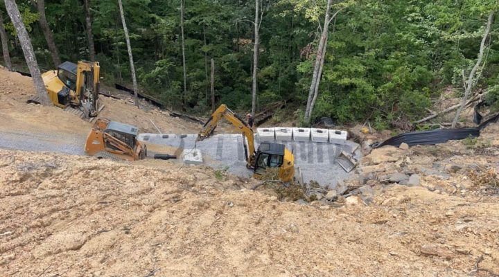 Multi-Machine Excavation and Foundation Work on a Steep Slope Three pieces of heavy equipment working simultaneously on a steep, wooded slope to install concrete foundation blocks for a new structure.