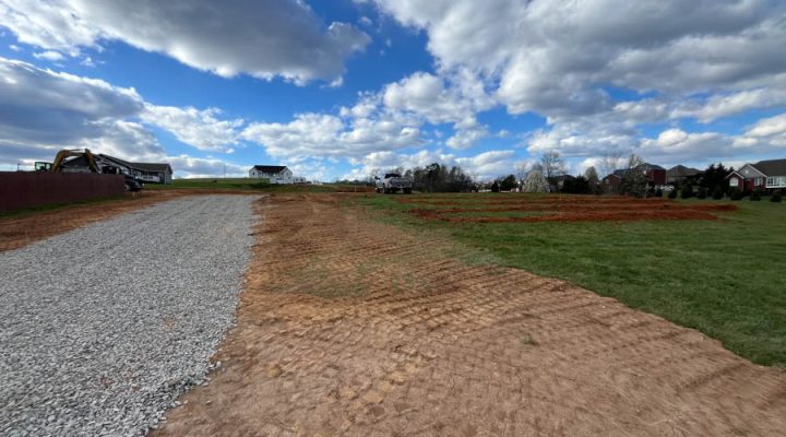 Gravel Driveway Installation and Site Grading for a New Home A newly installed gravel driveway running alongside a freshly graded, tire-tracked dirt area, with construction equipment and houses visible in the distance under a dramatic cloudy sky.