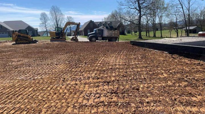Residential Lot Grading and Site Preparation in a Subdivision A freshly graded residential lot with visible tire tracks, featuring a skid steer, excavator, and dump truck near new homes under construction on a sunny day.