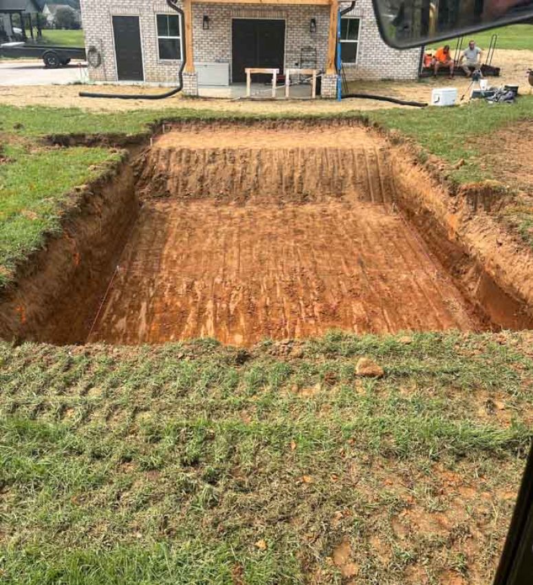 Precise Foundation Trenching for a New Custom Home A deep, rectangular excavation for a home’s foundation, with clean, straight walls and tire tracks on the compacted soil, located in front of a modern brick house.