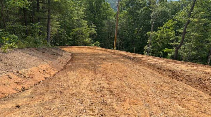 Newly Constructed Dirt Access Road Through a Wooded Area A winding, freshly graded dirt road cutting through a dense forest, showing clear tire tracks and a view of distant mountains under a partly cloudy sky.