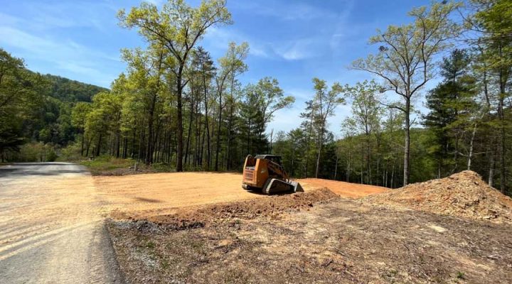 Skid Steer Leveling Soil on a Forested Building Site An orange skid steer loader working on a cleared, flat building pad at the edge of a wooded area, with a pile of excavated earth nearby under a clear sky.