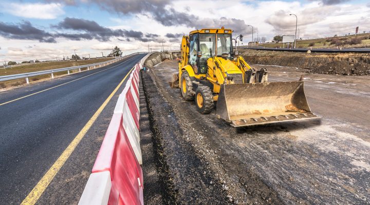 Commercial Roadway and Infrastructure Project A yellow front-end loader working on a highway construction site next to a newly paved road, with red and white safety barriers in place.