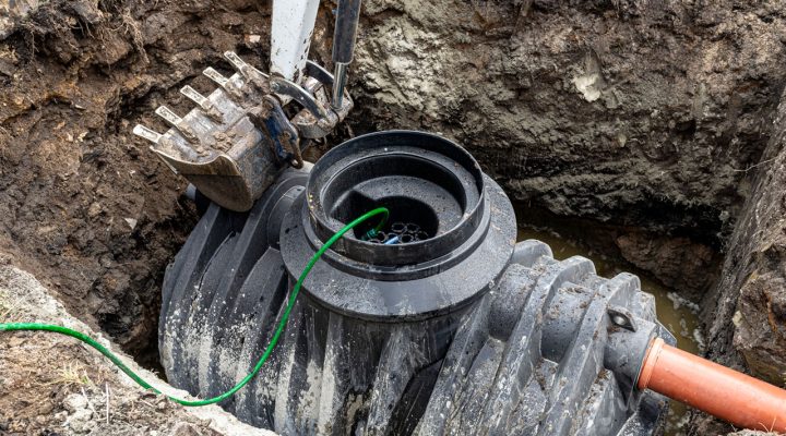 Underground Septic System Component Installation Close-up view of a black plastic septic tank component being installed in a muddy trench, with an excavator bucket positioned above.